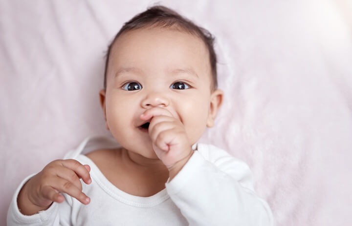 Smiling baby lying on a pink blanket with fingers in their mouth.