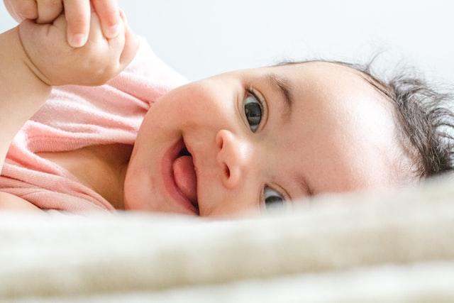Smiling baby lying on a bed while holding an adult's finger.