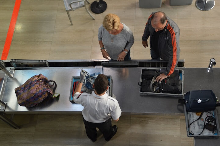 Airport security officer inspecting carry-on bags at a screening checkpoint, viewed from above.