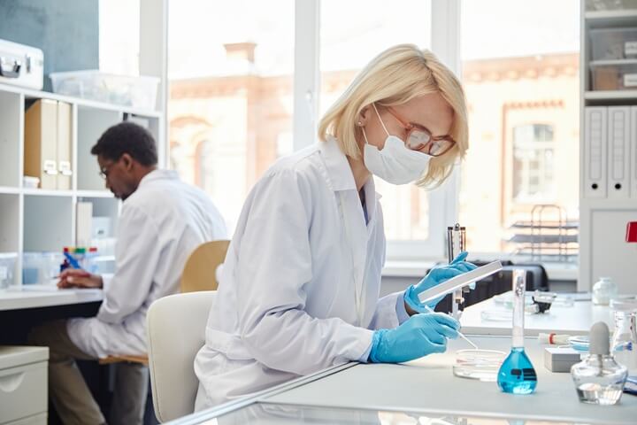 Lab technician wearing a mask and gloves working with petri dishes and glassware.
