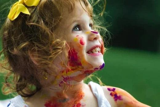 Young child outdoors with colorful paint smeared across their face and shoulder.